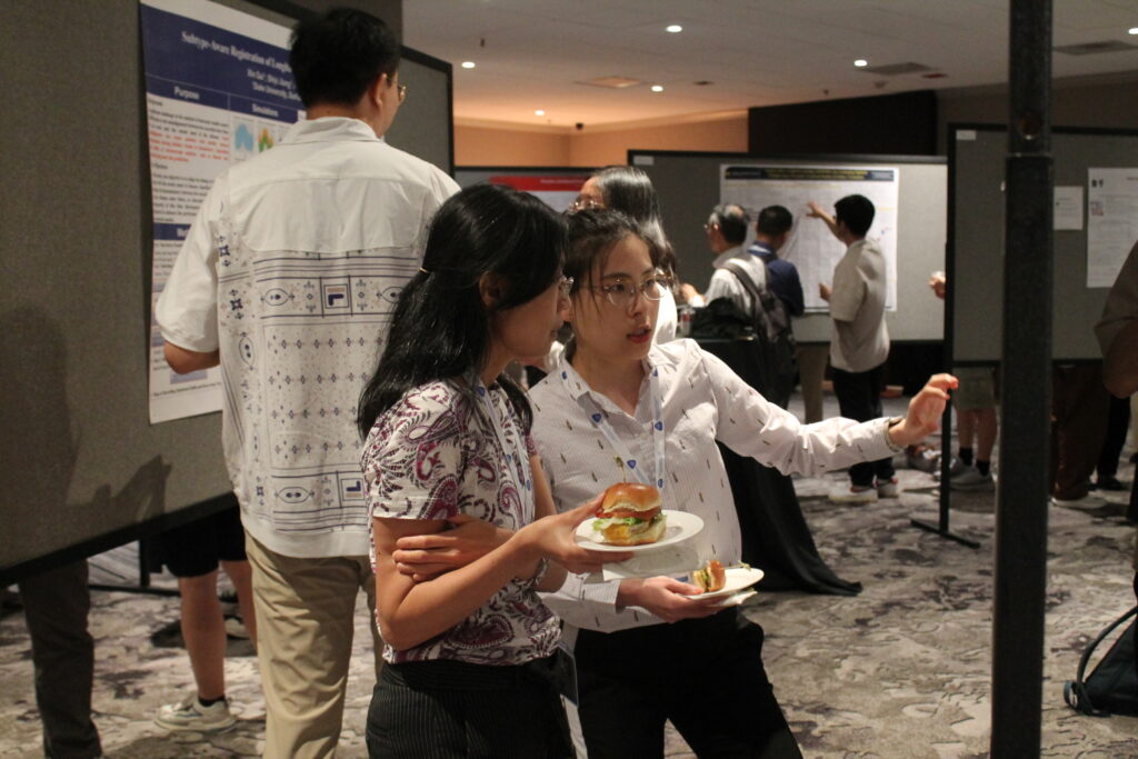 Two women holding food discuss a poster while other participants mingle in the background