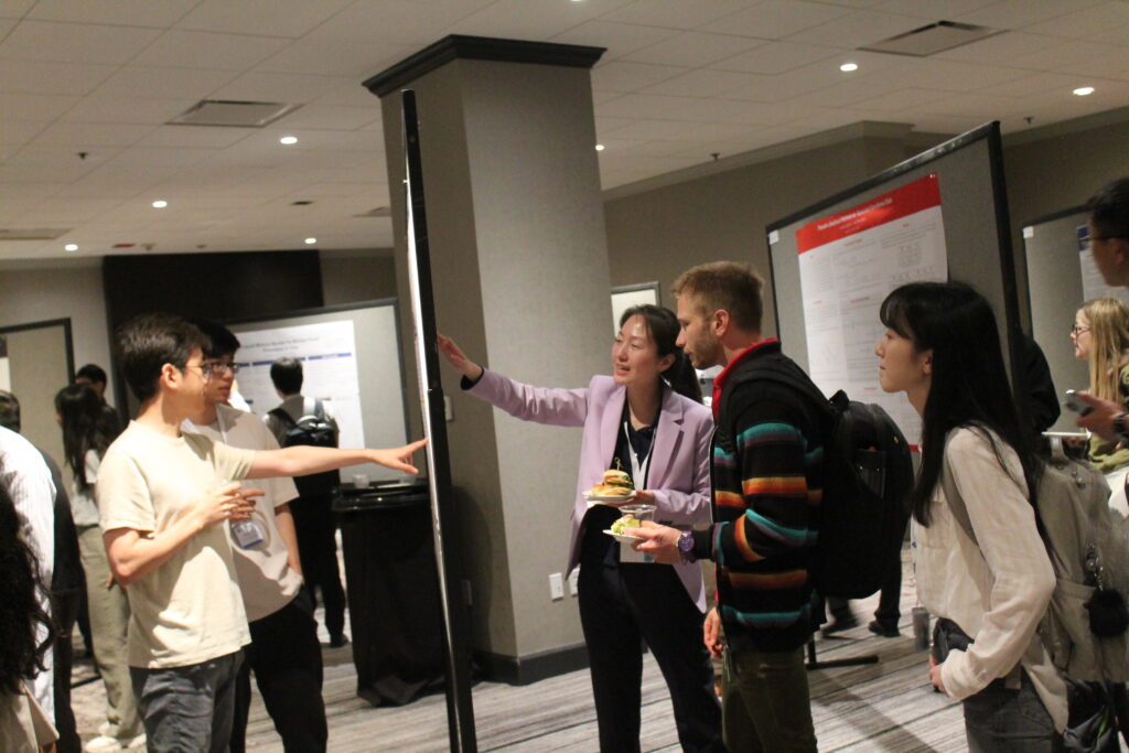 Two poster presenters (man in white shirt, woman in lavender blazer) point to their work on opposite sides of a poster board while other attendees look and listen.